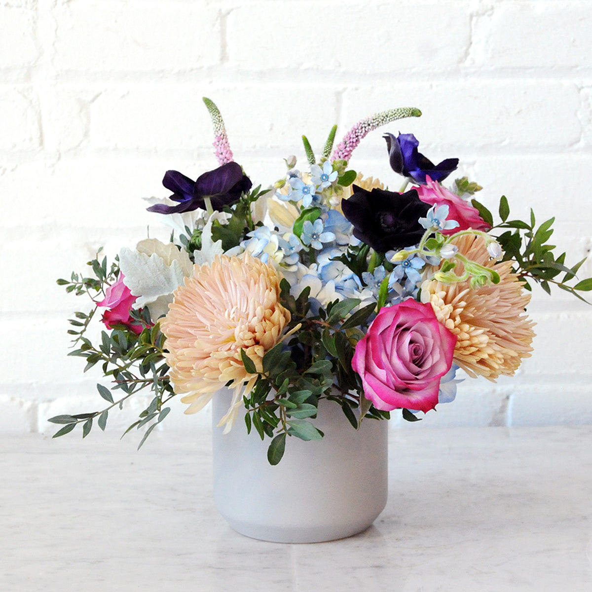 Spruce flowers Emerson arrangement shown in a gray vase with textural foliage silver dusty miller leaves and Lilac roses with cremons
