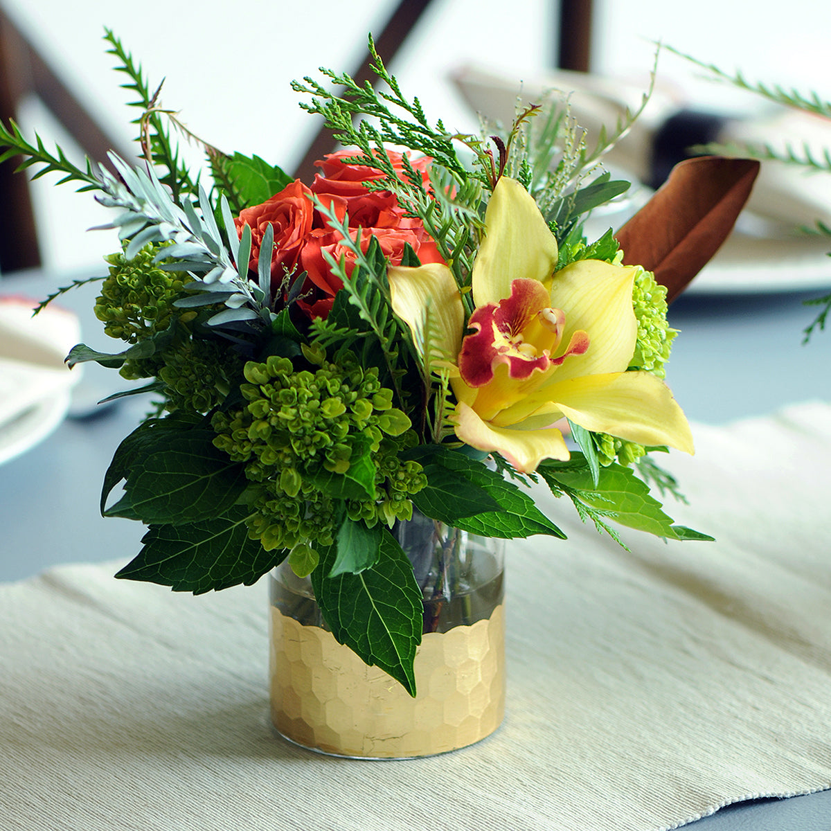 Floral arrangement with red roses and yellow orchids in a clear and gold vase on a table.