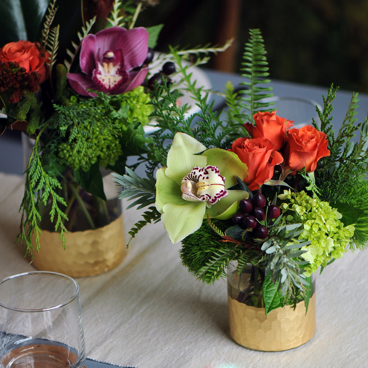 Two floral arrangements in gold vases on a table with a glass of water.