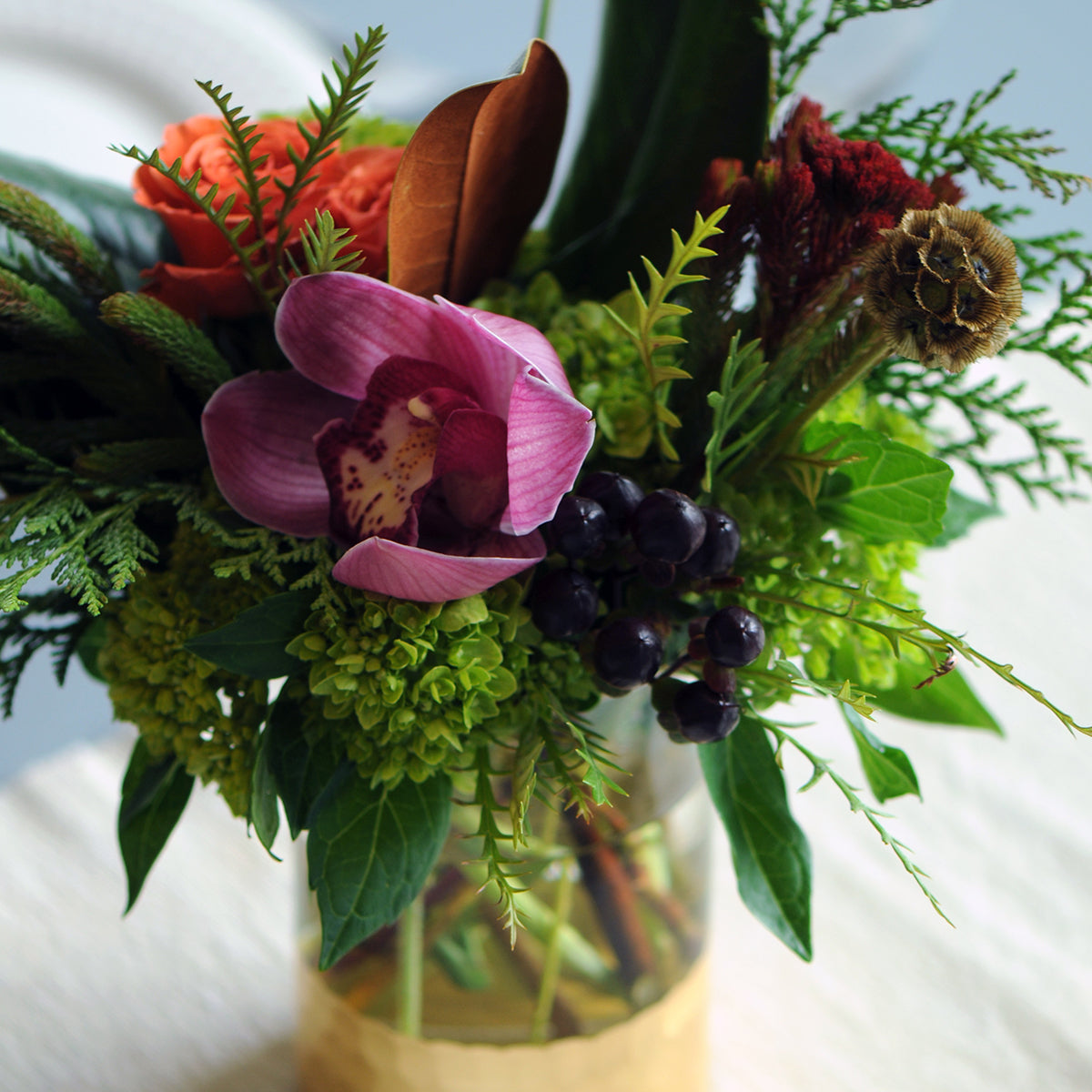 Bouquet of flowers with a pink orchid, green leaves, and purple berries in a clear vase.