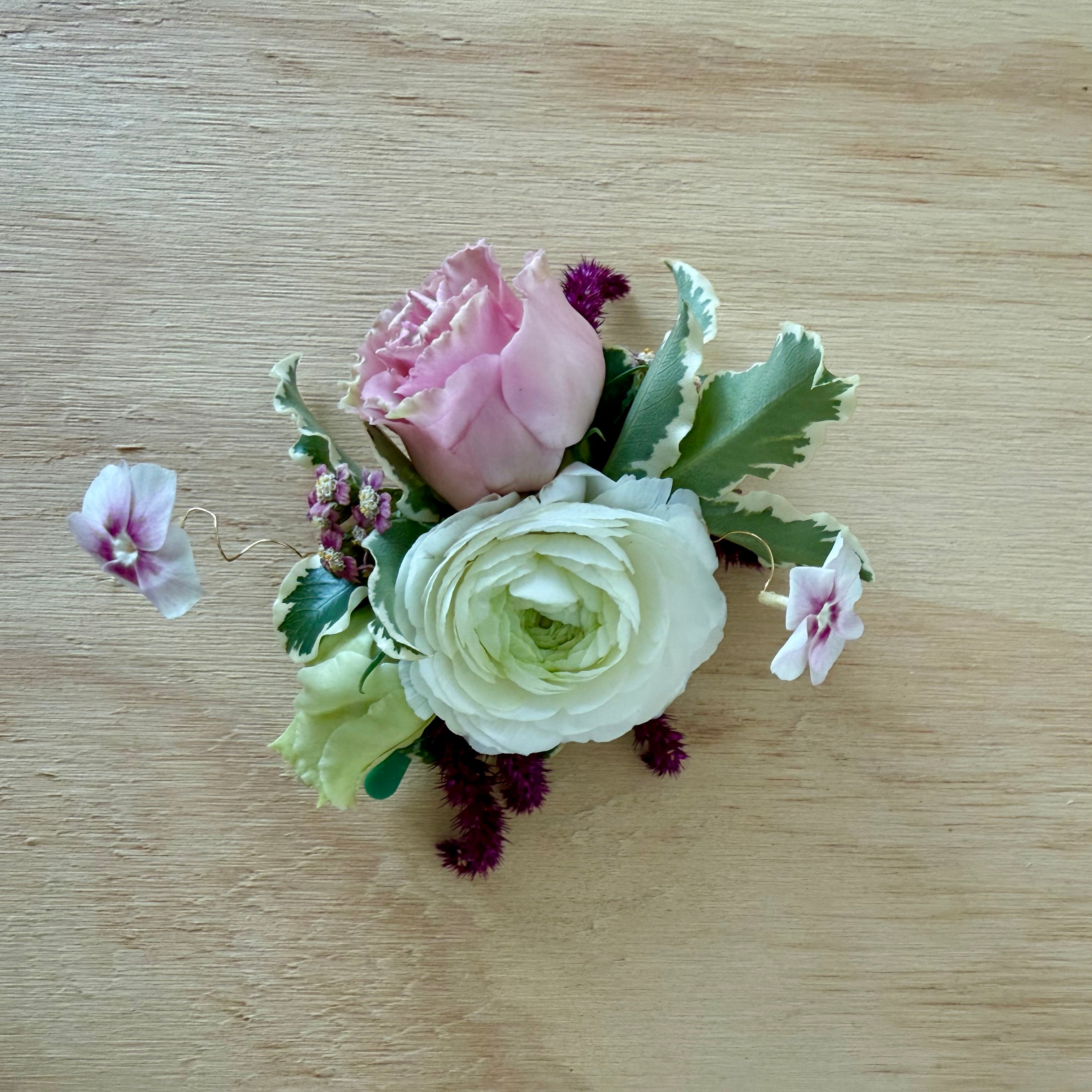 A small floral corsage or boutonniere arrangement featuring a pink rose, a white ranunculus, green leaves, and small purple accents, placed on a light wood surface.