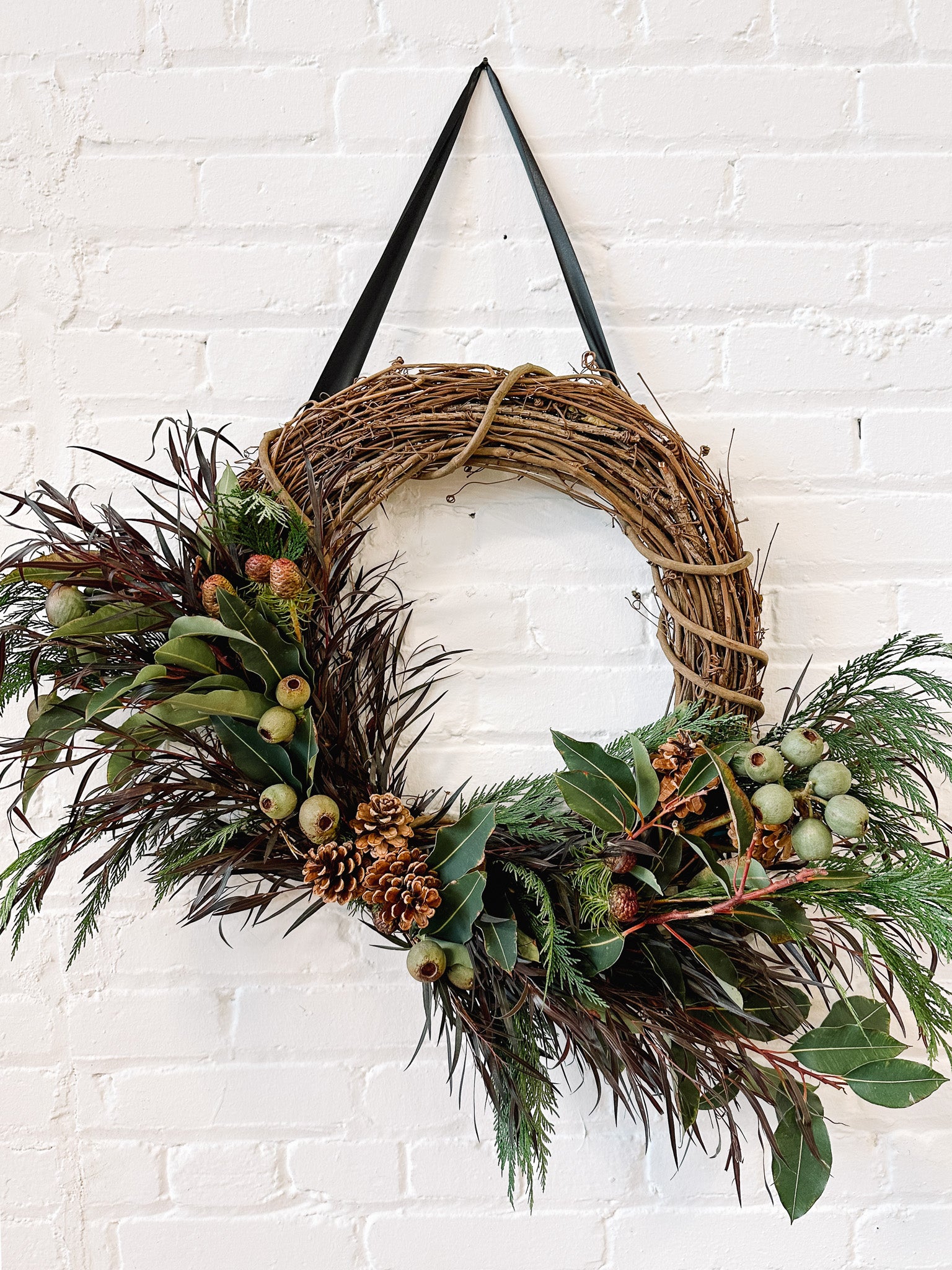 A festive wreath made with a mix of fresh and dried elements, including green foliages, pine cones, and berries, displayed against a white brick wall.