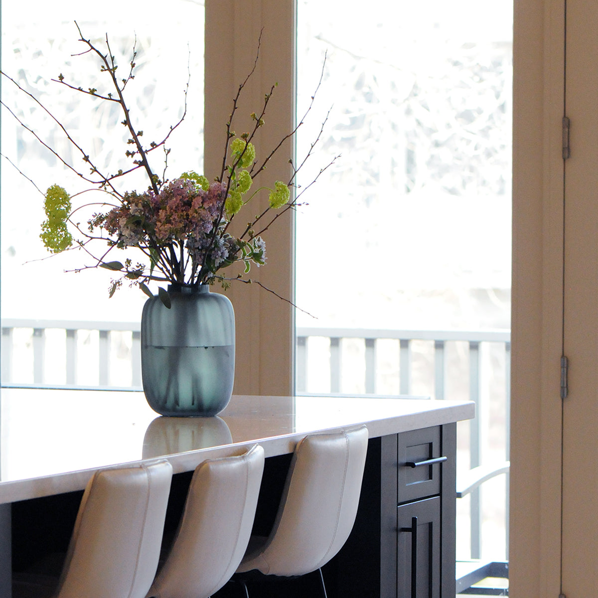 Vase with flowers on a kitchen counter with white chairs and a window in the background