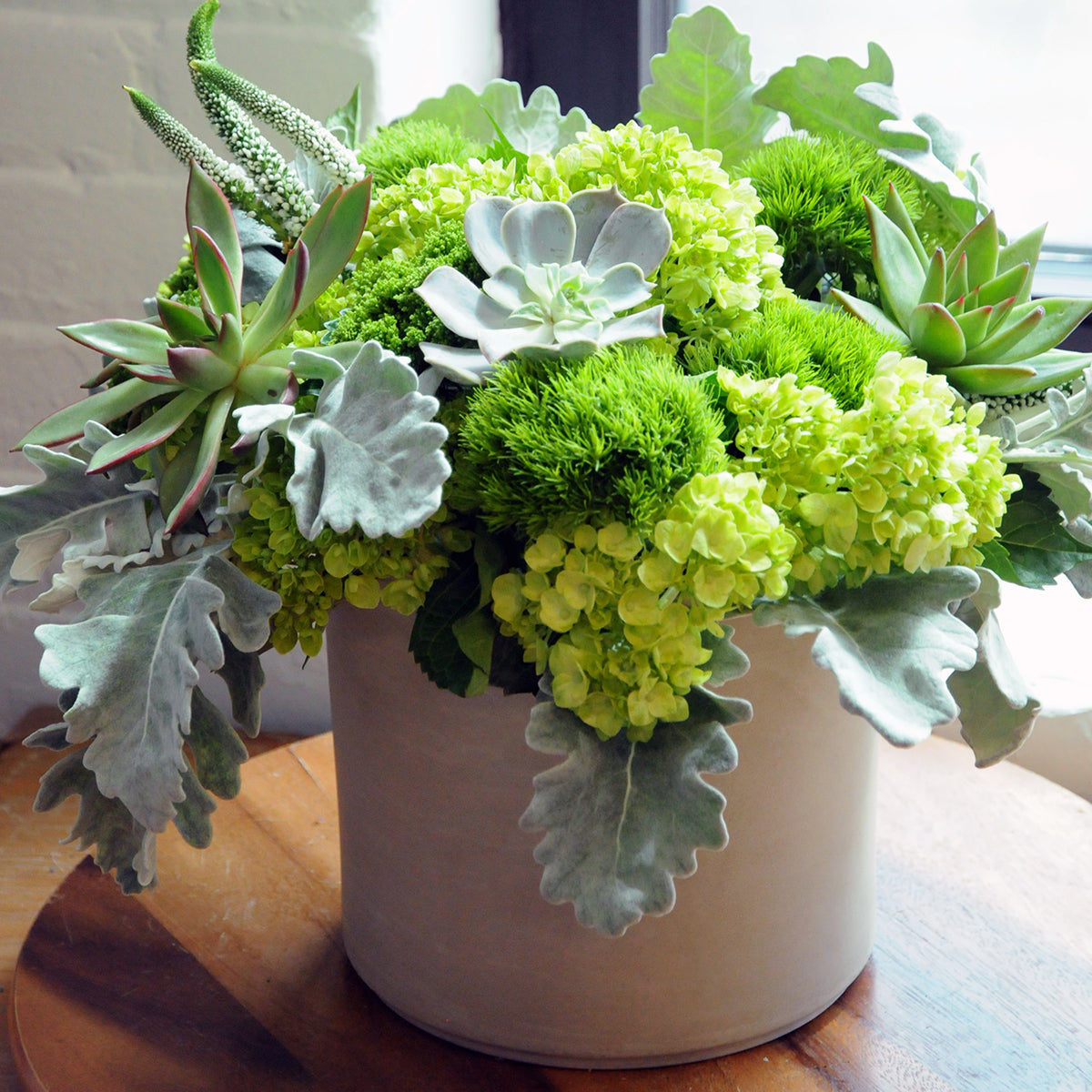 Floral arrangement from Spruce Flowers  with green succulents and monochromatic green flowers in a cream vase on a wooden surface
