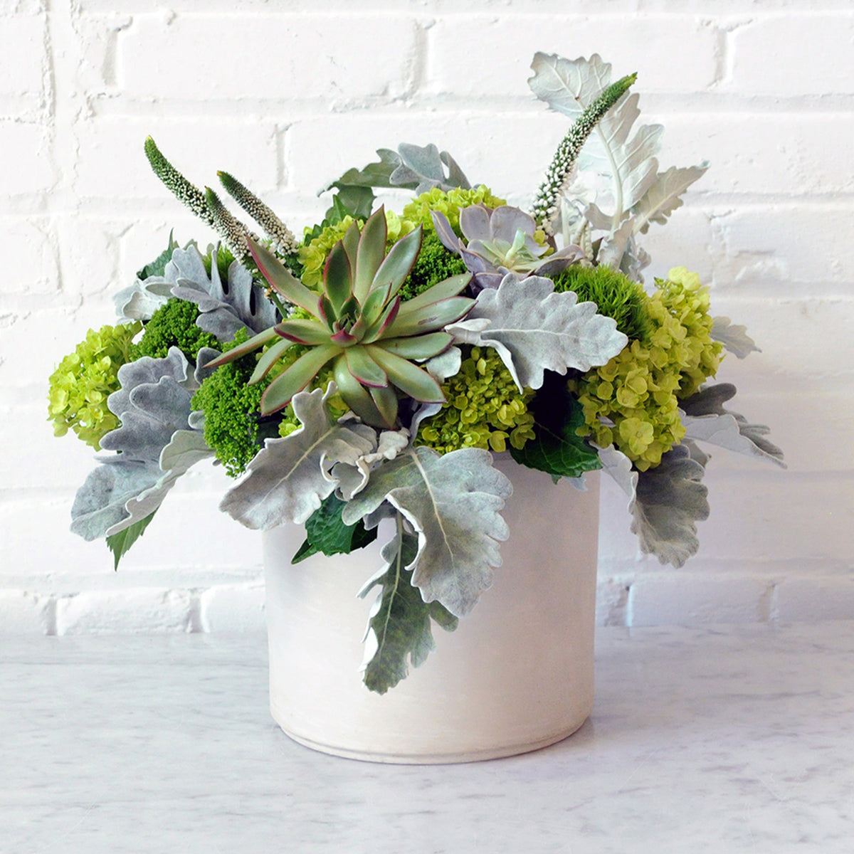 Decorative arrangement of succulents and greenery in a white pot against a white brick wall.