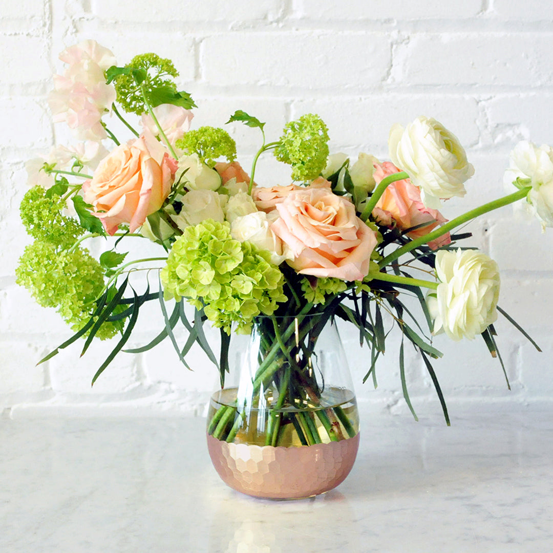 A floral arrangement featuring peach roses, white spray roses, and green viburnum in an Elsa kettle vase with gold detailing on the base, placed against a white brick background.