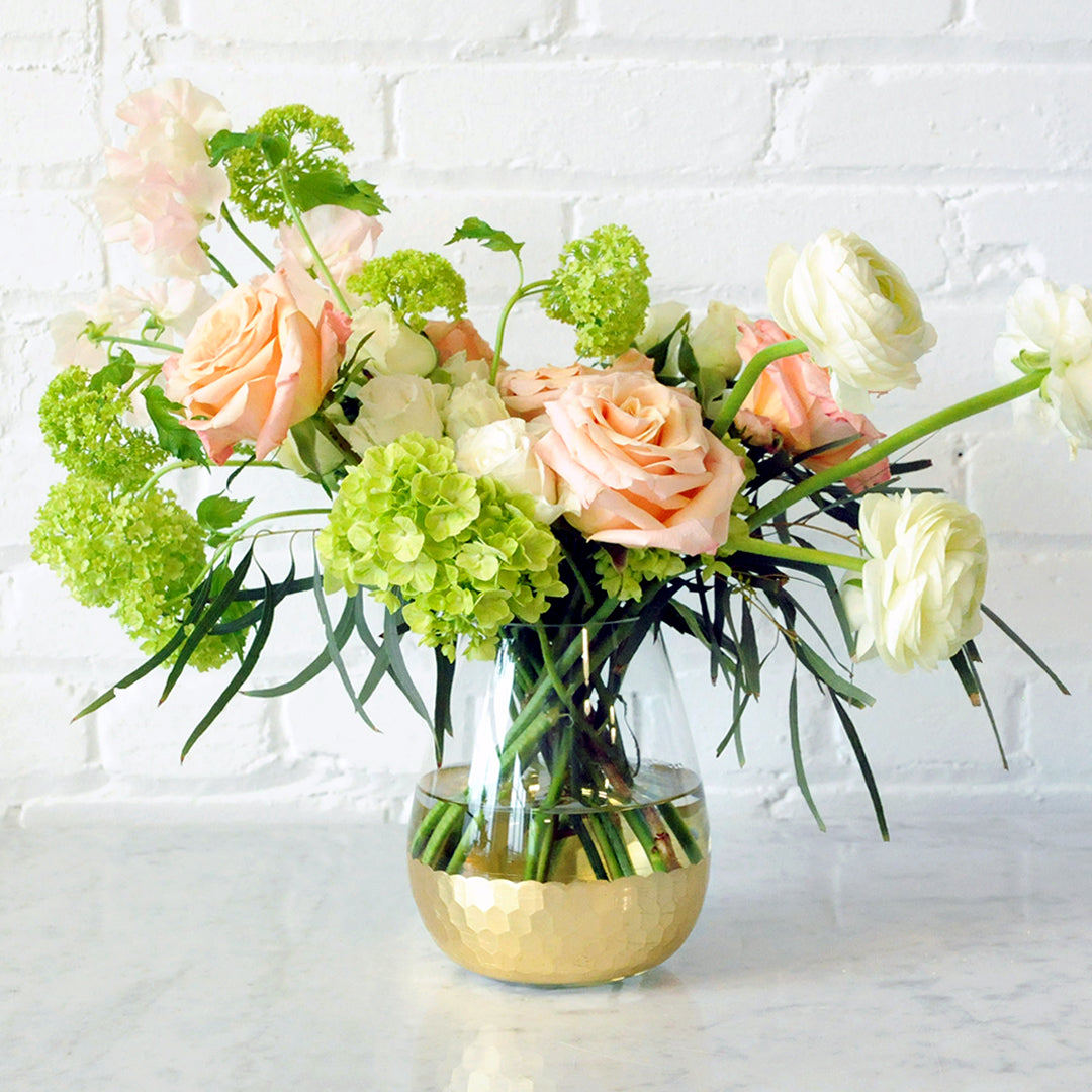 Image of Floral arrangement on table. A circular vase with textured gold finish to the base. The floral arrangement is whimsical and asymmetric with green Hydrangea Spray roses and peach rose accents with dainty Sweet Pea and Ranunculus accents