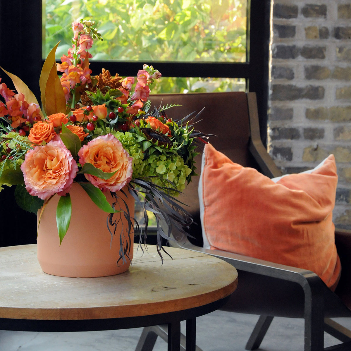 Spruce flowers Marshall floral arrangement in fall tones with textural foliages set on a coffee table with a chair and cushion.