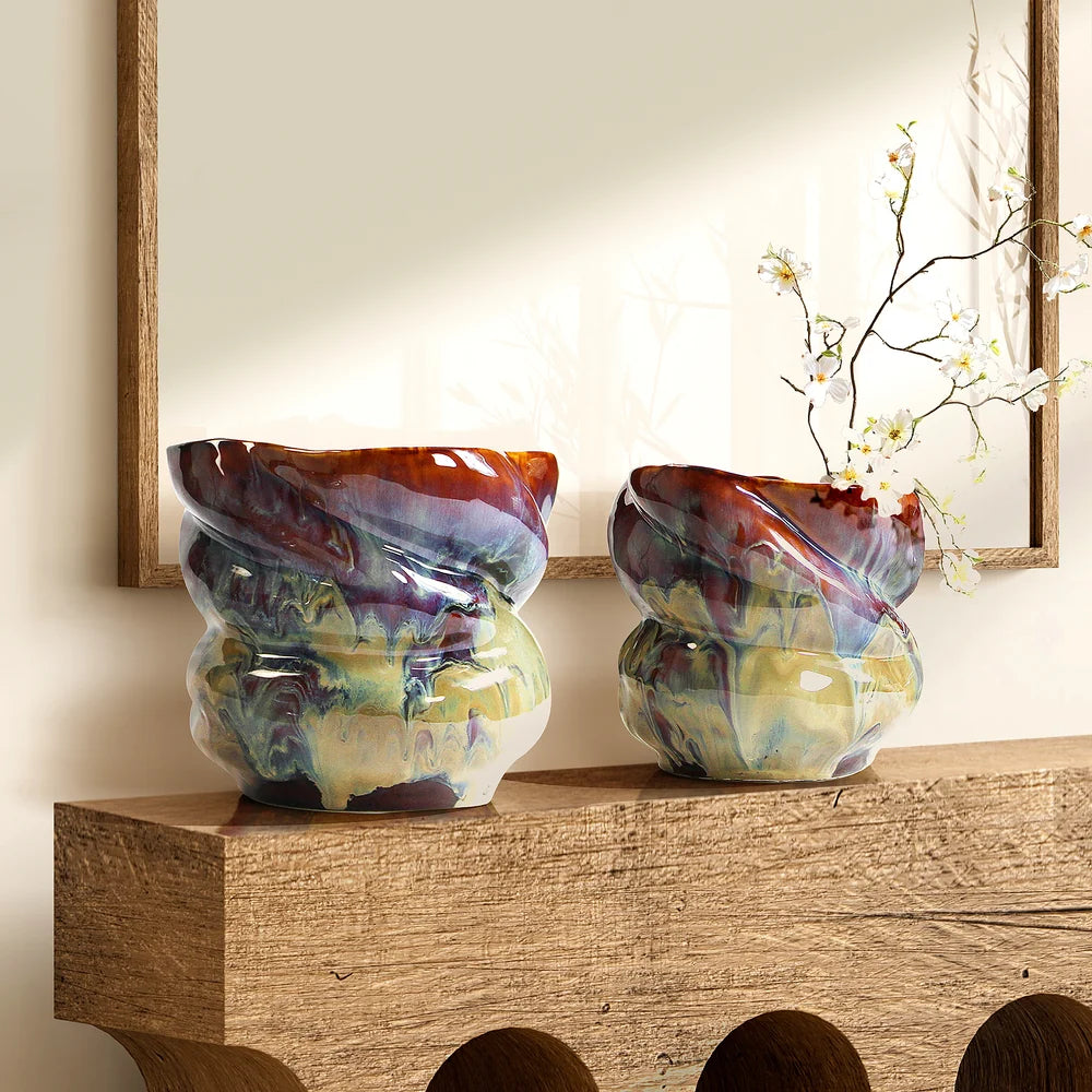 Two ceramic vases with a glossy, marbled multicolor glaze in shades of purple, green, yellow, and brown, displayed on a rustic wooden mantelpiece with a mirror and dried branches in the background.