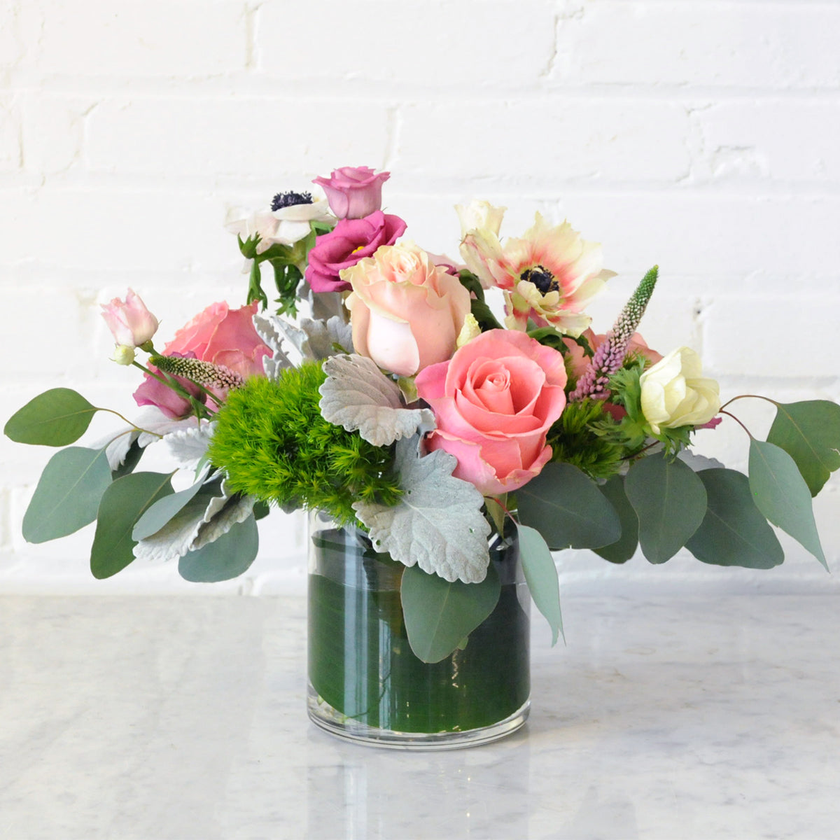 Spruce Flowers Montrose arrangement in a glass cylinder vase complete with a leaf wrap. The arrangement has mixed shades of pink roses Trick Dianthus, silver $ Eucalyptus and Dusty Miller foliage.