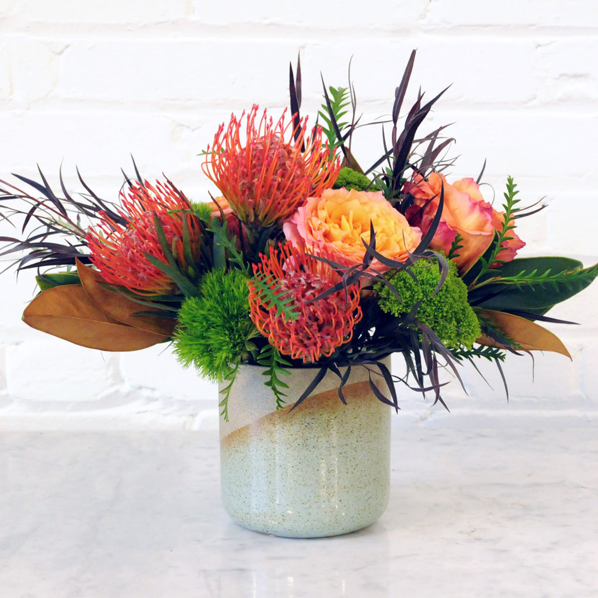 Photo of floral arrangement showing Rich tones of Red and Orange for this fun festive fall arrangement with textural foliage in a Celeste Marbella Vase