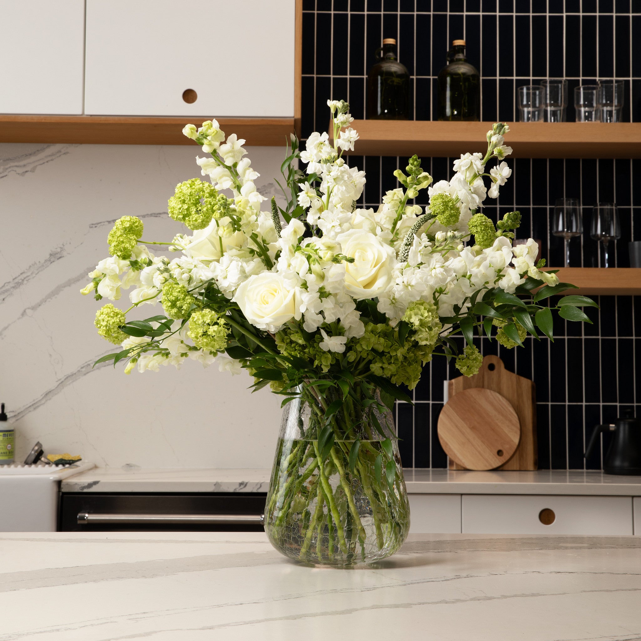 A large floral arrangement of white roses, white stock, and green hydrangea blooms in a clear glass vase, set on a light marble kitchen countertop with a modern kitchen background.