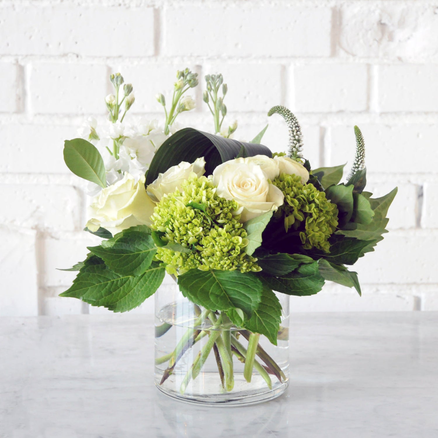 White Florals with green hydrangea in a clear glass cylinder vase. Image is an example of the styling rather than the exact arrangement that will be sent