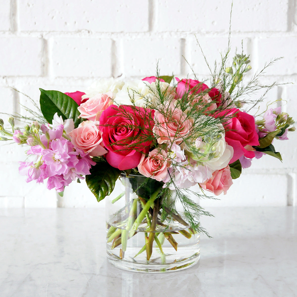 Spruce Flowers Elsie arrangement shown in a glass vase Pink Flloyd Roses Scented pink stock and pink spray roses artfully arranged on a bed of white Hydrangea.