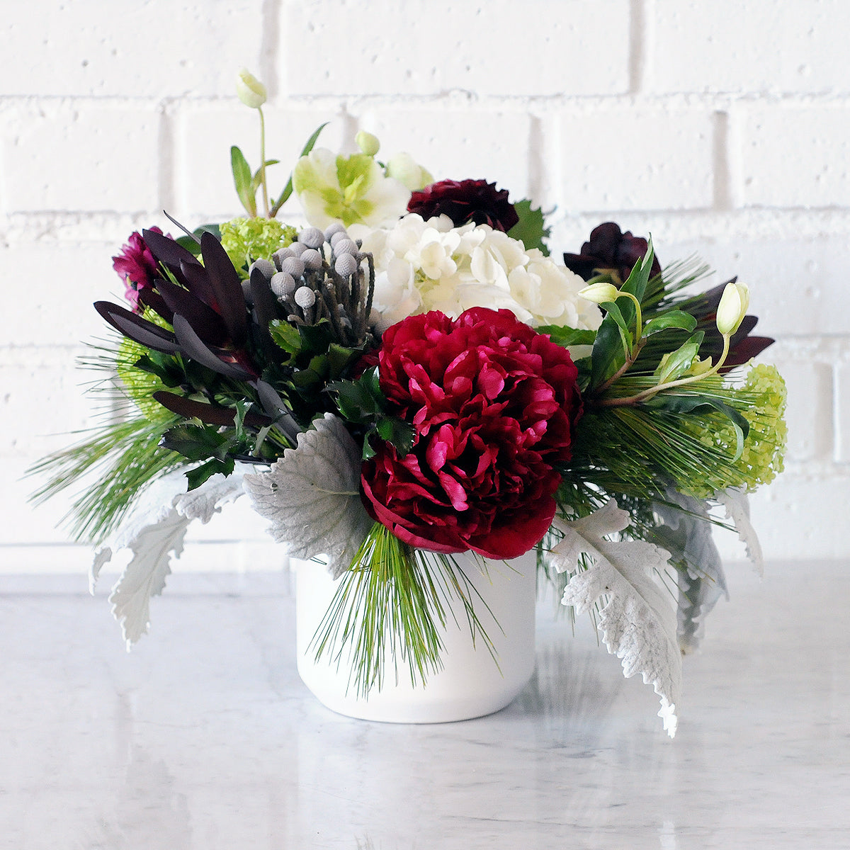 Front view of Hawthorne arrangement in white ceramic vase featuring bold red peonies and textural florals