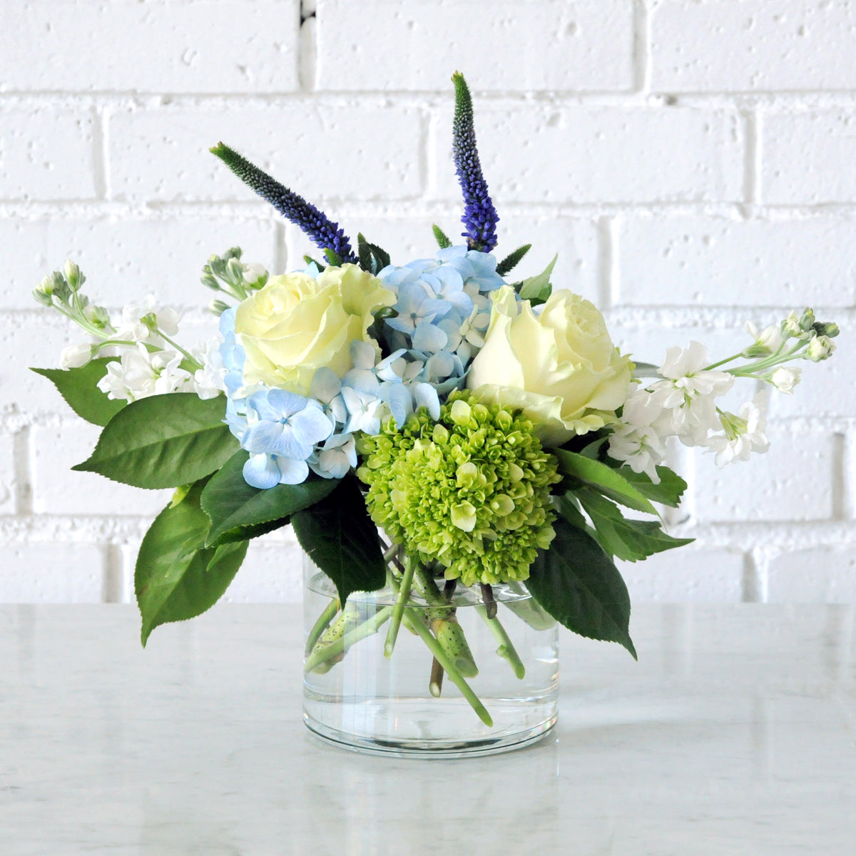 Spruce Flowers arrangement of Blue hydrangea green nini greens white roses and stock in a clear glass vase