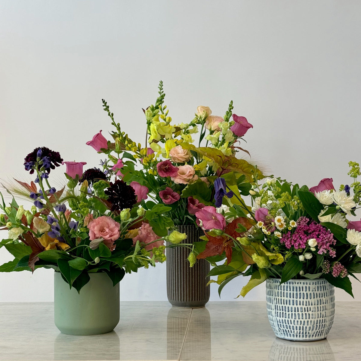 Three flower arrangements in different vases on a light surface with a neutral background