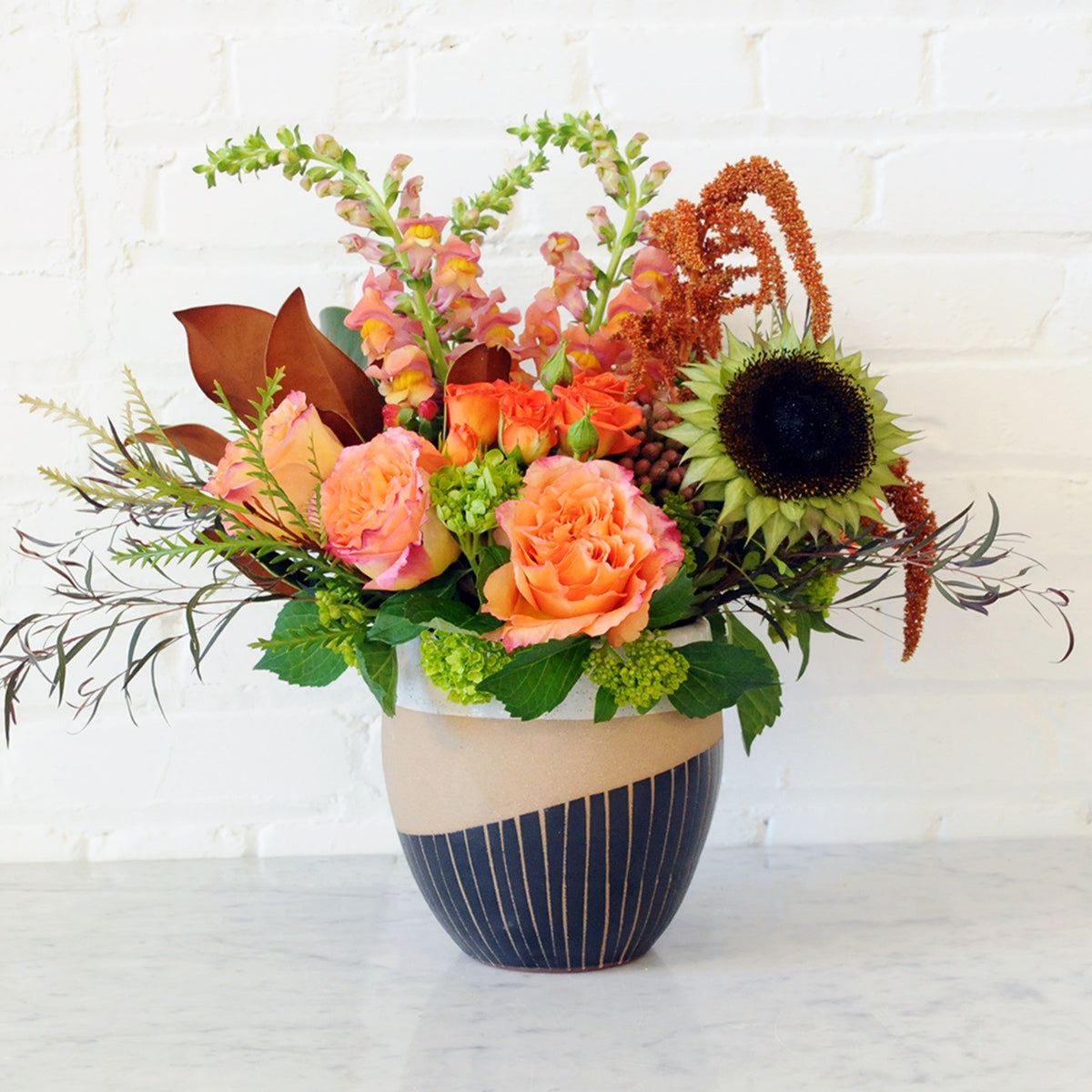 A vibrant autumnal floral arrangement in a ceramic vase, featuring orange roses, a sunflower, and green foliage, set against a white brick wall background.