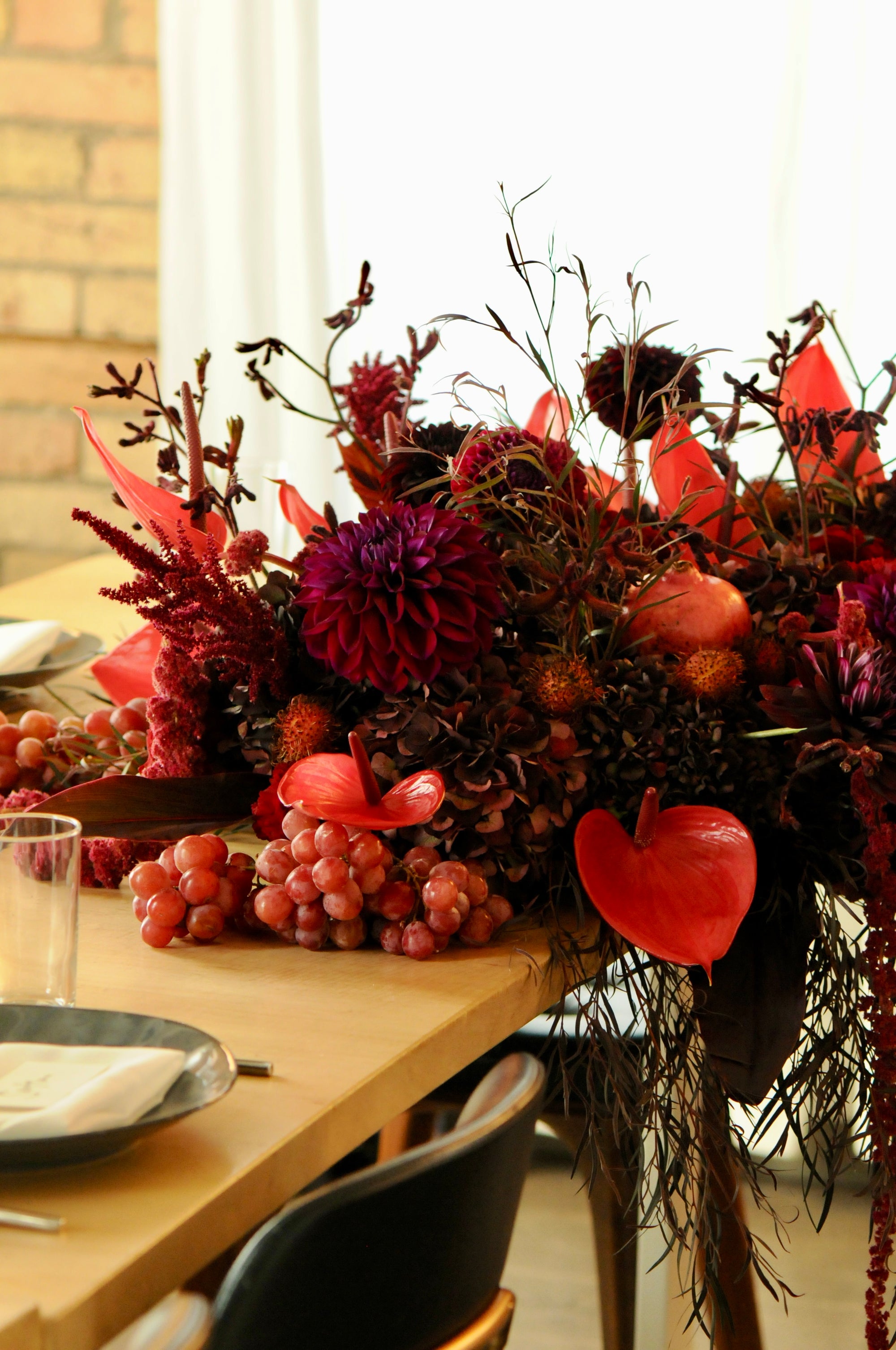 An autumnal floral centerpiece arranged on a wooden dining table, featuring deep red and burgundy flowers, clusters of red grapes, and warm orange accents in a styled indoor setting.