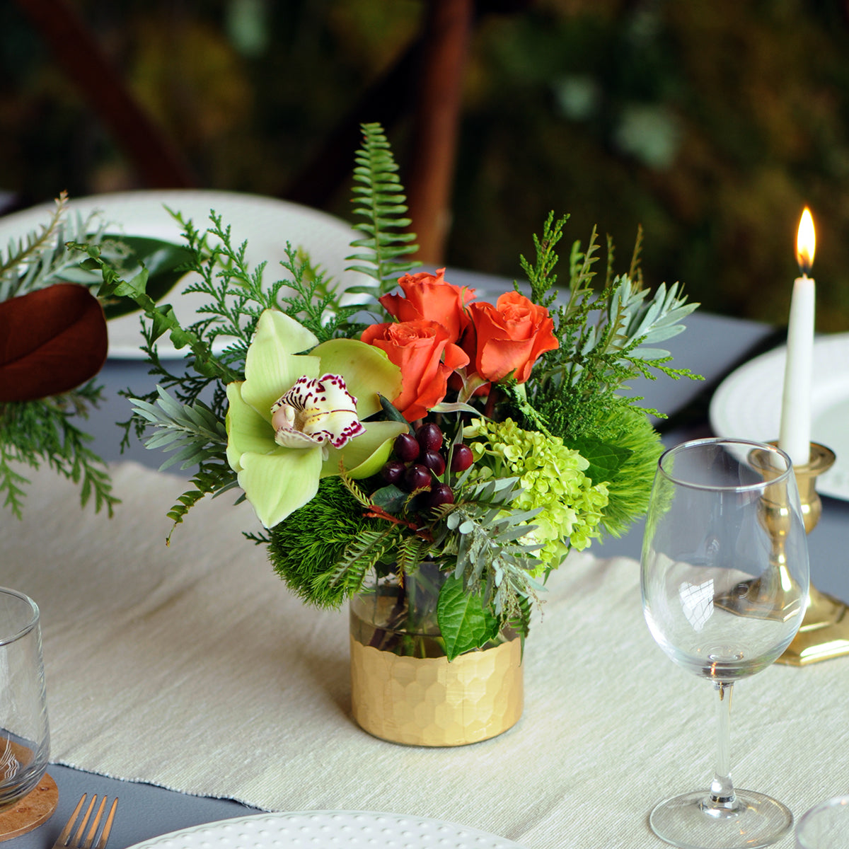 Decorative floral arrangement with red roses and greenery on a table setting with glasses and a candle.