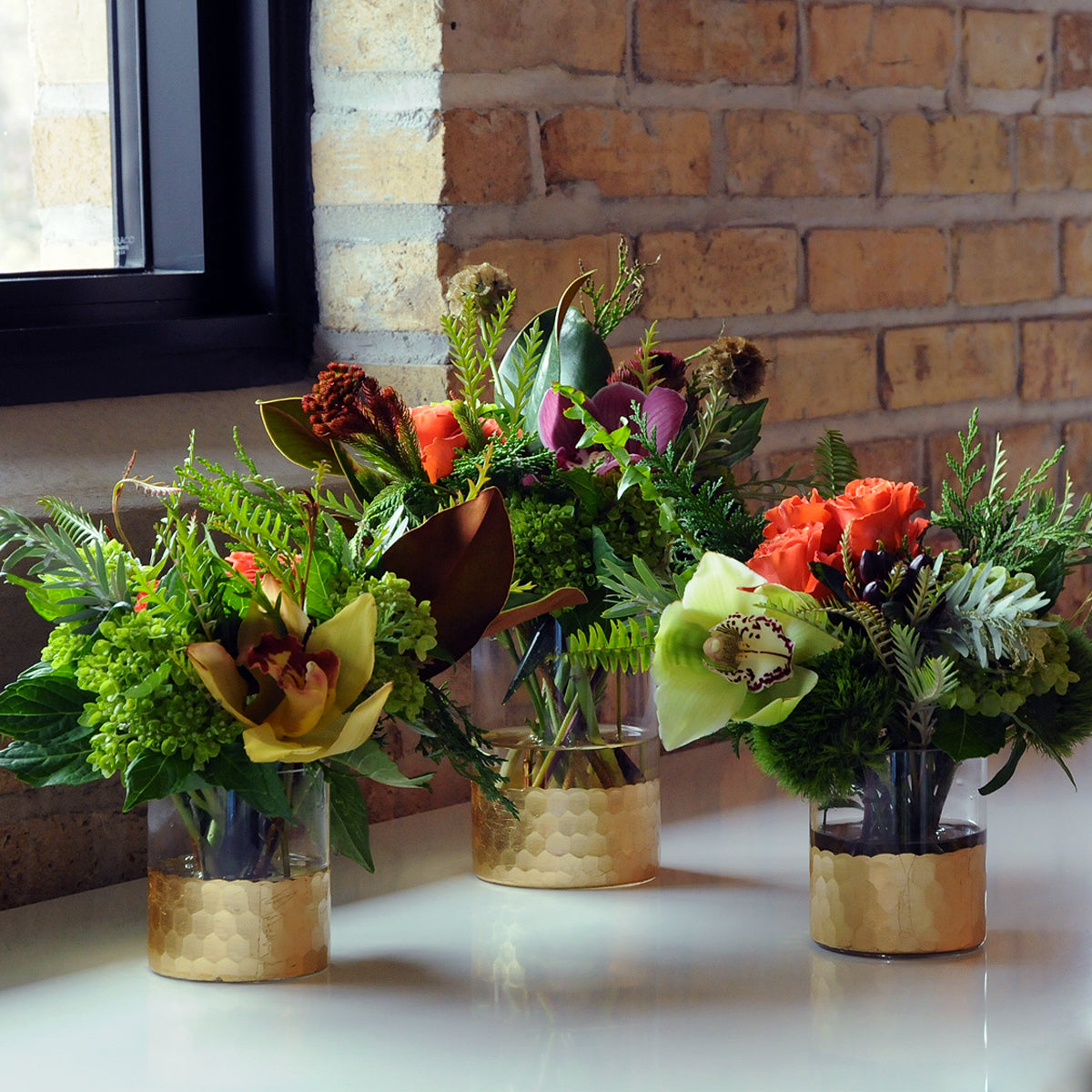 Three floral arrangements in gold vases on a white surface with a brick wall background.