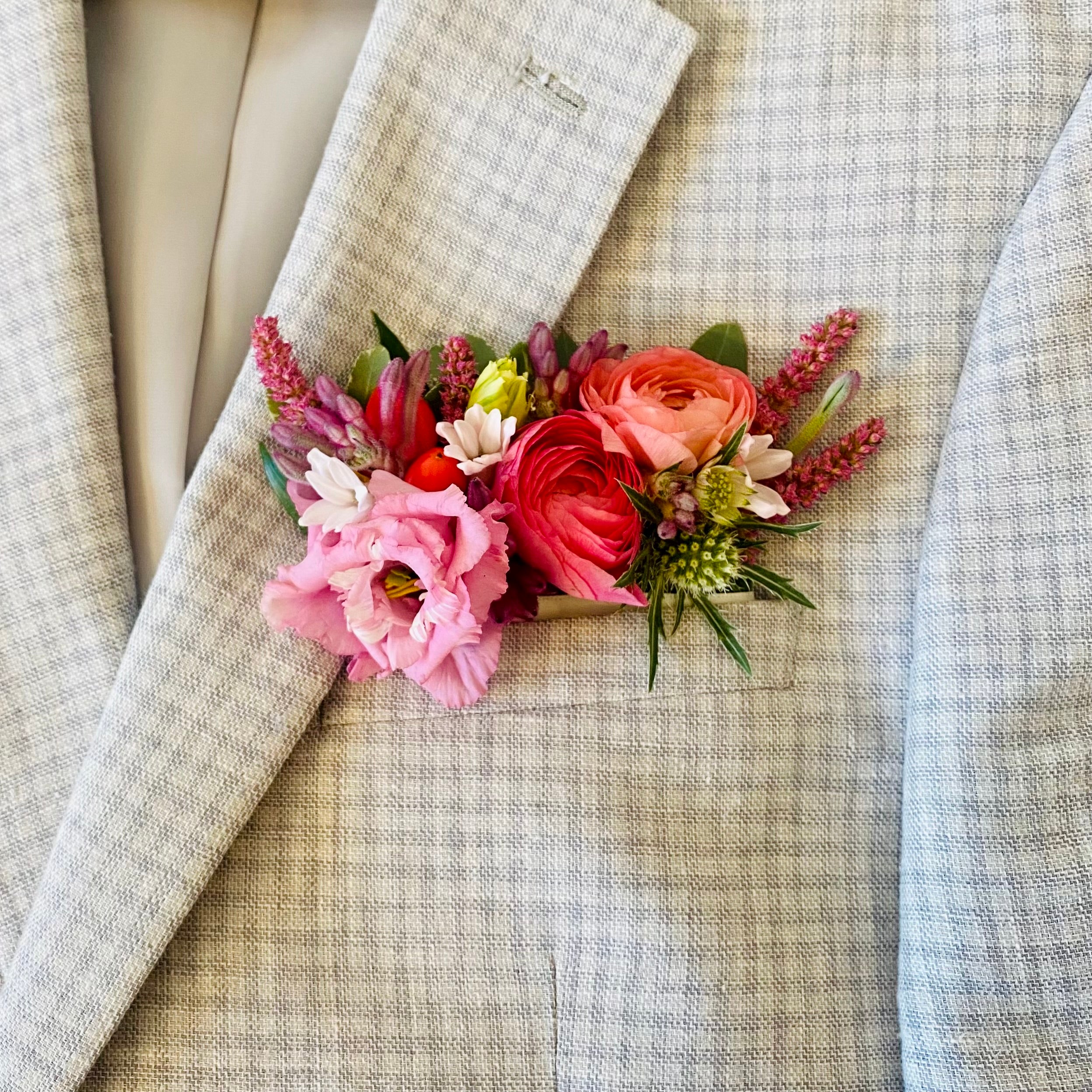 A close-up of a colorful floral Pocket Tuck pinned to a light beige and gray plaid suit jacket. The flower arrangement features pink, red, and peach ranunculus s with green foliage and small white and yellow accents.