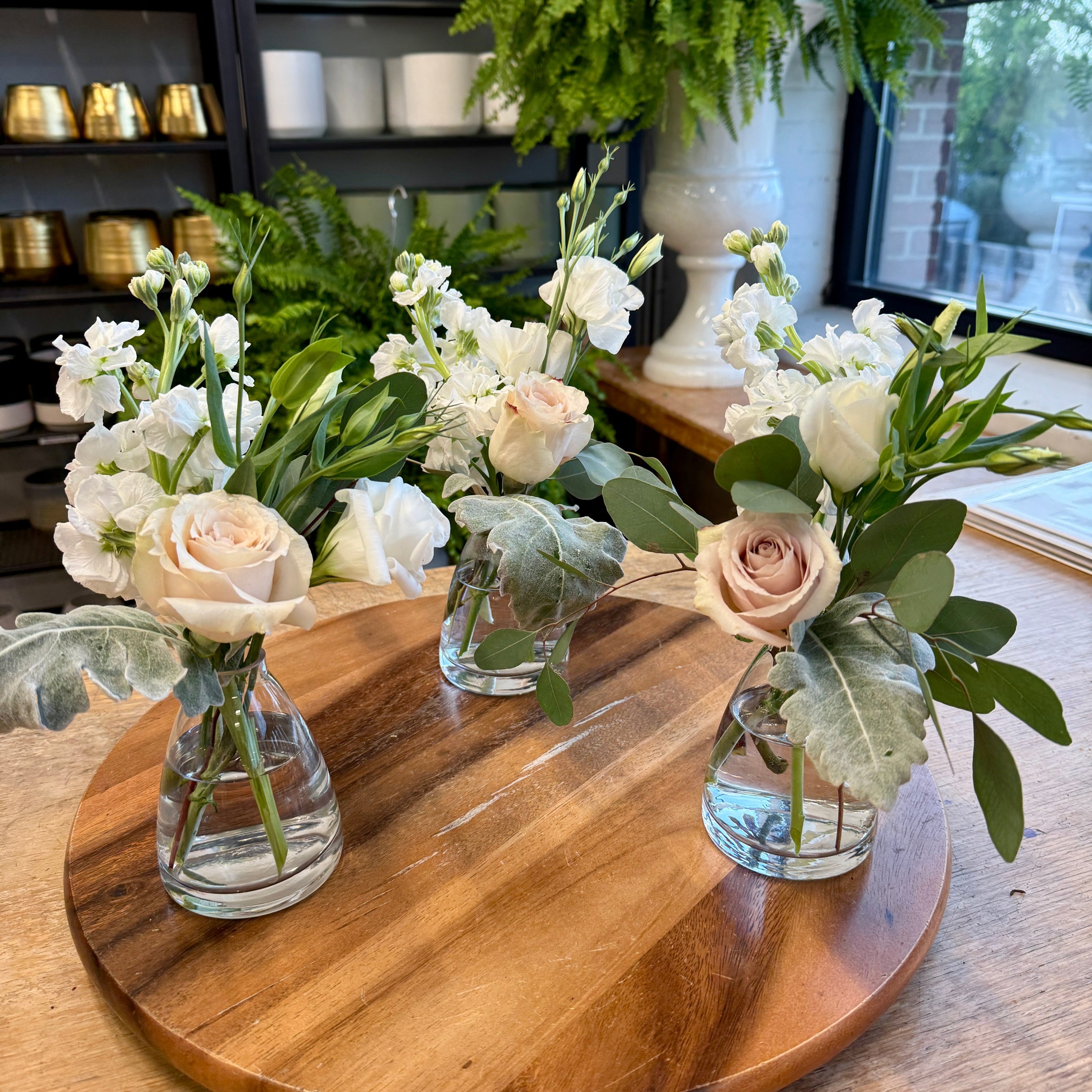 Three small floral arrangements in clear glass vials with blush pink roses, white flowers, and green foliage, displayed on a round wooden board in a shop setting.