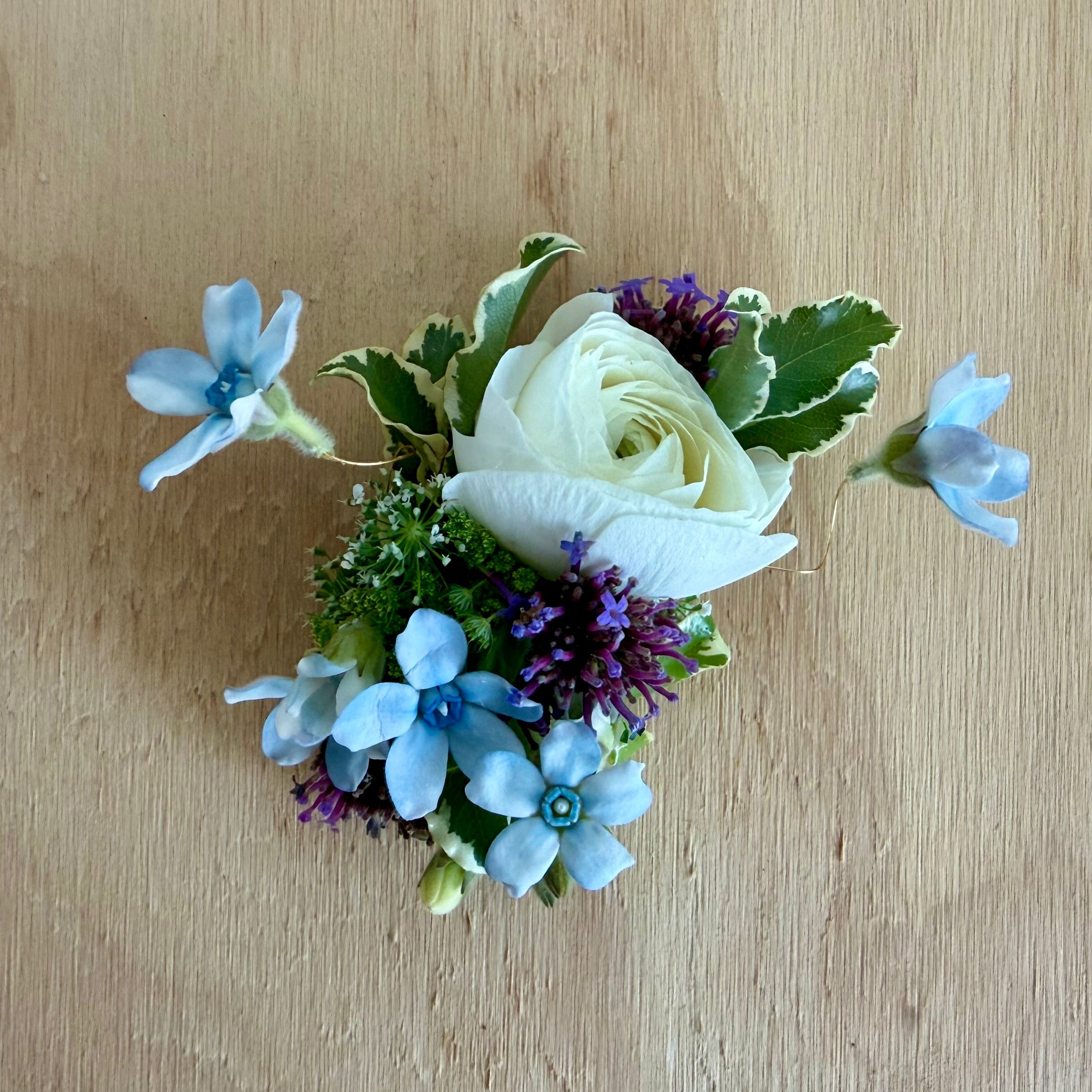 A small floral boutonniere featuring a white rose, light blue starflowers, purple filler blooms, and green leaves, arranged on a light wood background.