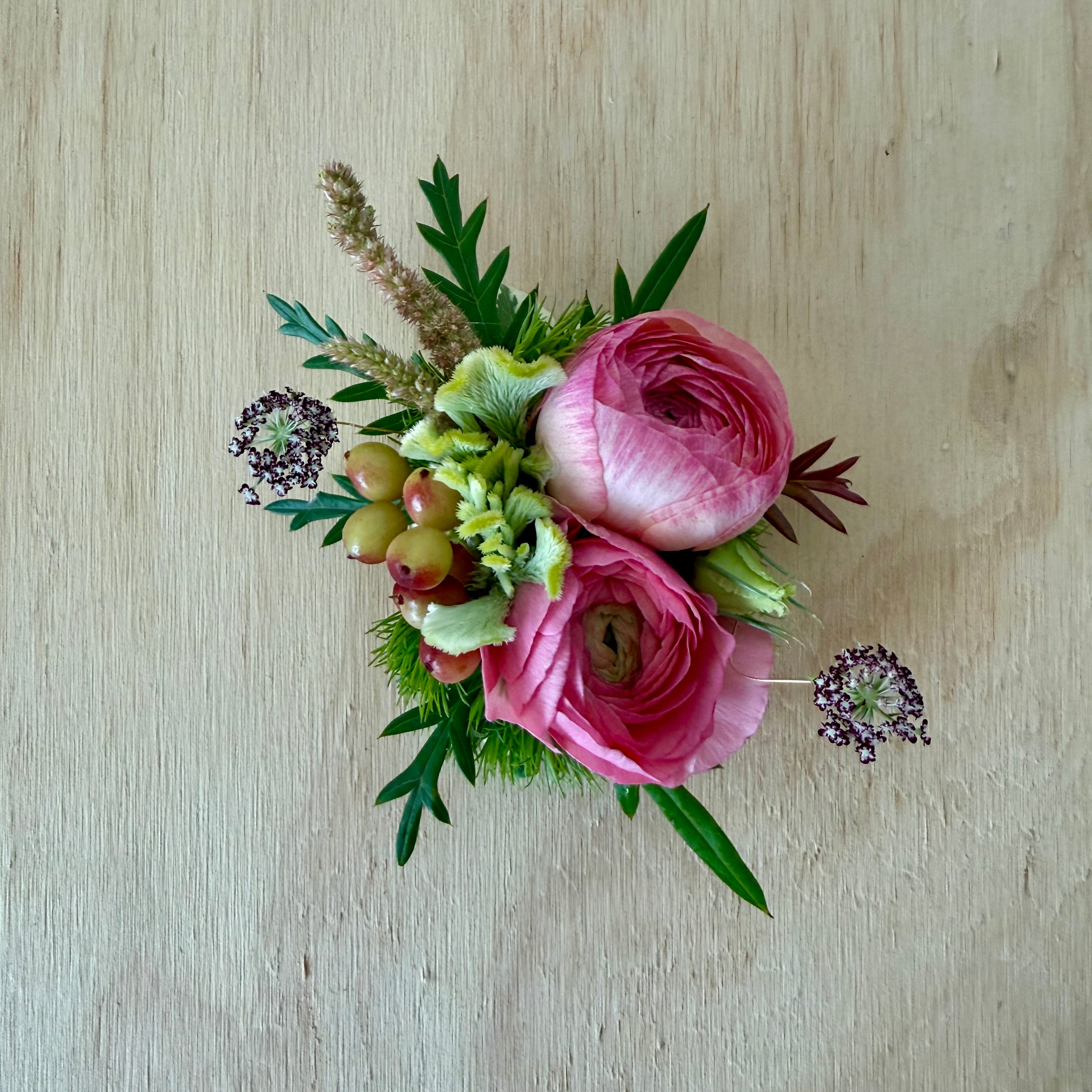 A small wearable corsage arrangement with two pink ranunculus flowers, green foliage, and a few pale green berries, photographed from above on a light wood surface.