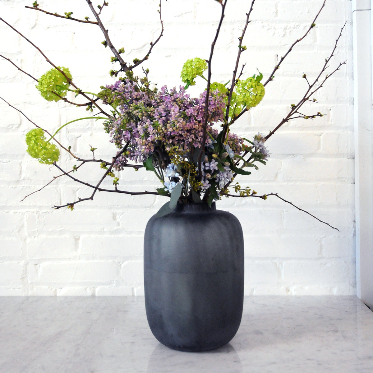Dark gray vase with a bouquet of flowers and branches against a white brick wall.