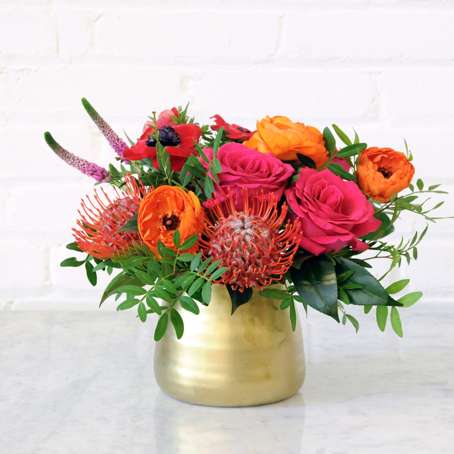 Front photo or a gold tegan ceramic vase with Spruce Flowers Lexington Arrangement featuring Pink Floyd roses and orange Ranunculus.  