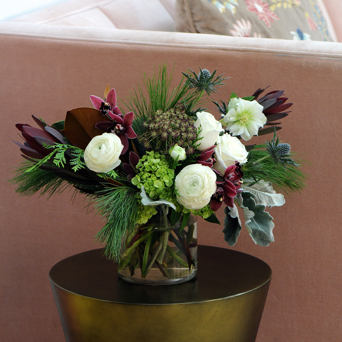 Bouquet of flowers in a clear vase on a round  table with a pink couch in the background.