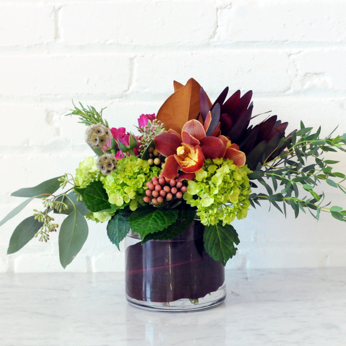 Colorful floral arrangement in a leaf wrapped vase against a white brick wall.