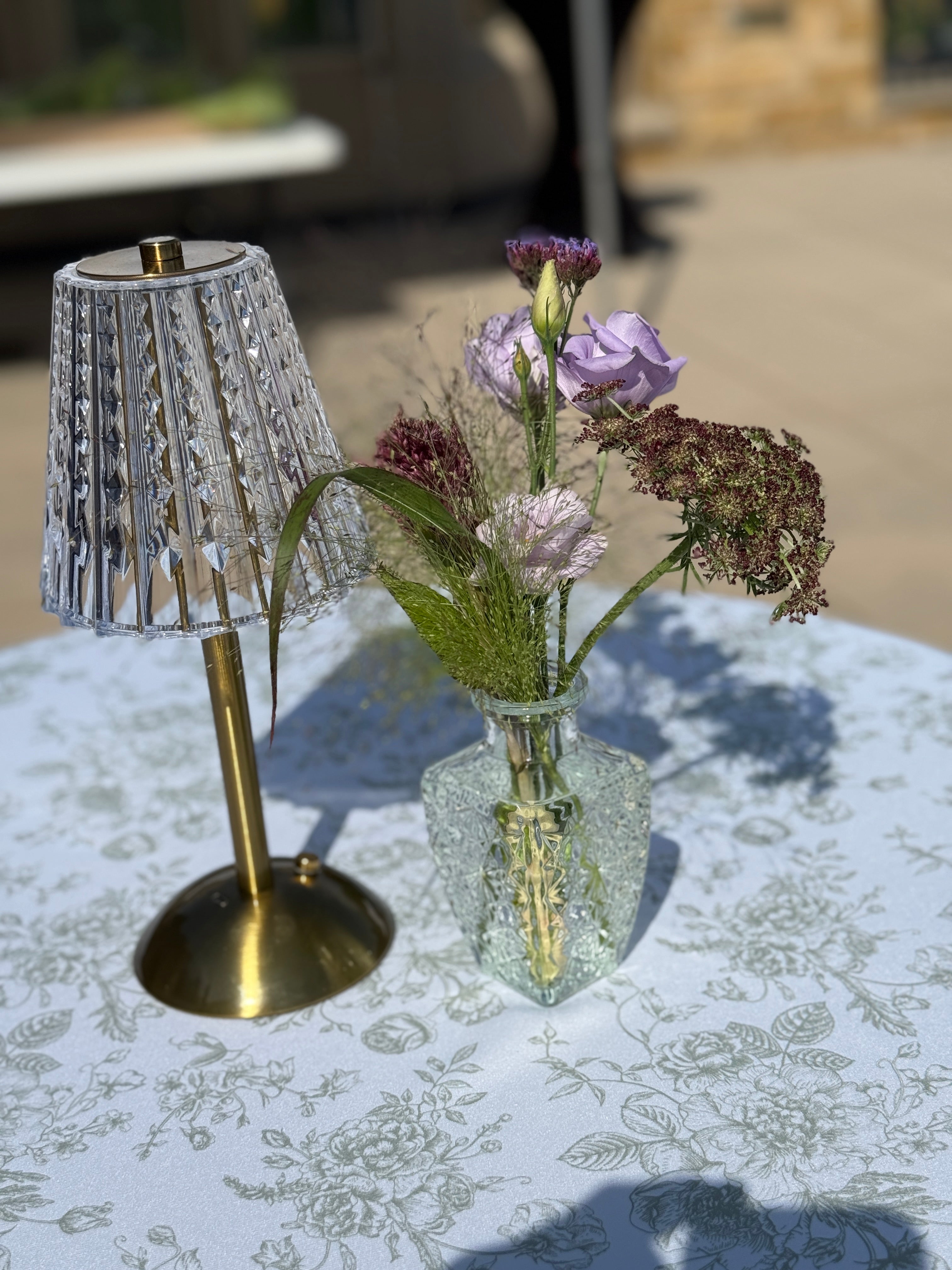 A small table lamp with a faceted crystal-textured shade and gold base sits beside a clear glass vase holding purple and pink flowers, set on a white floral tablecloth outdoors.