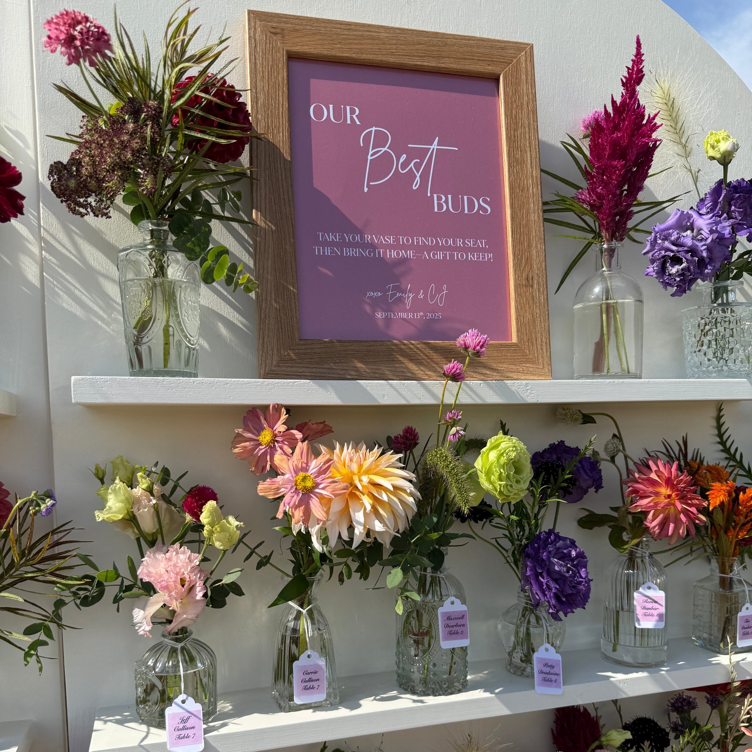 A floral display on white shelves with multiple clear glass vases holding colorful flowers, including dahlias, roses, and ornamental grasses. A framed sign reading 'Our Best Buddies' hangs above the arrangement against a white wall.
