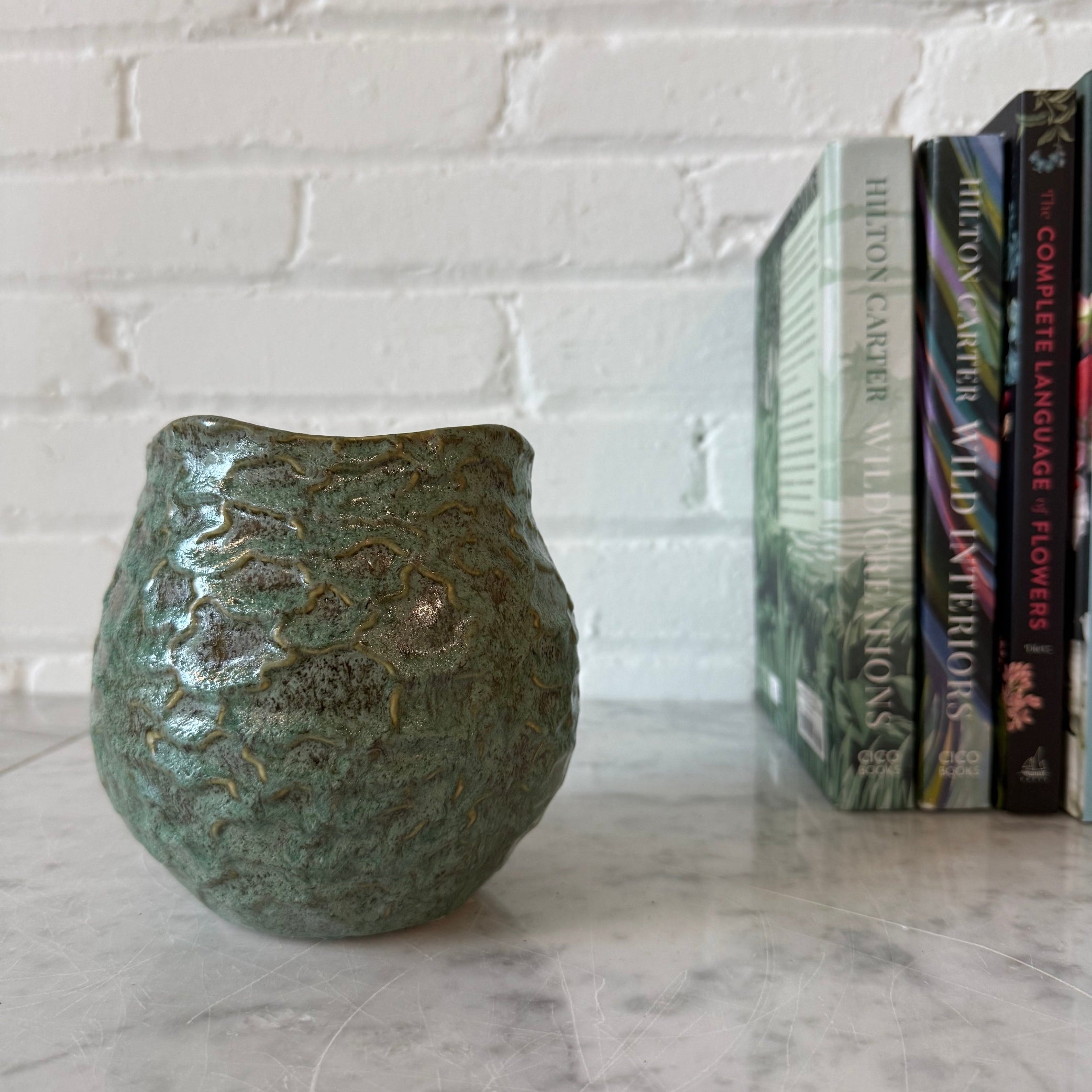 A small rounded ceramic vase with a textured, glossy green glaze sits on a light marble surface against a white painted brick wall, with a row of books standing upright in the background.