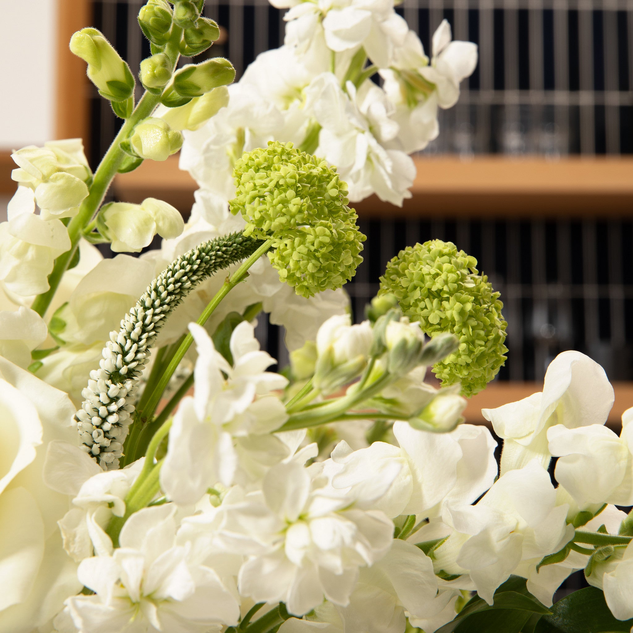 A close-up of a floral arrangement featuring white flowers and two prominent light green Viburnum blooms, with a green textured Veronica flowers set against a blurred indoor background.