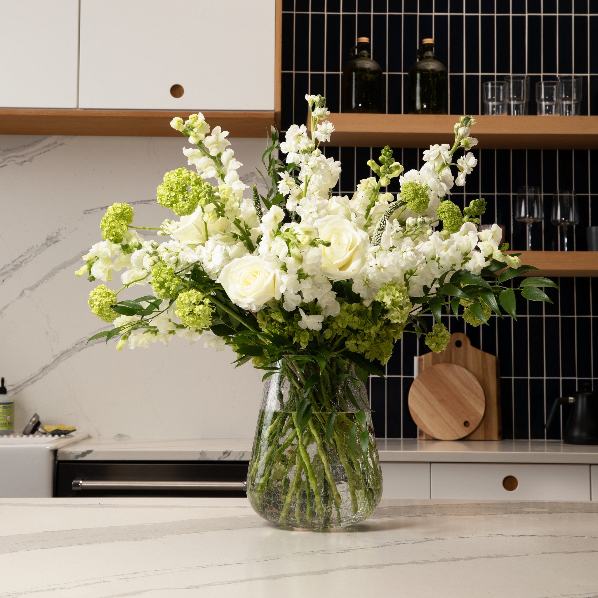 A large flower bouquet featuring white roses, white stock, and green hydrangea blooms in a crackle  glass vase, set on a kitchen countertop with a modern kitchen background.