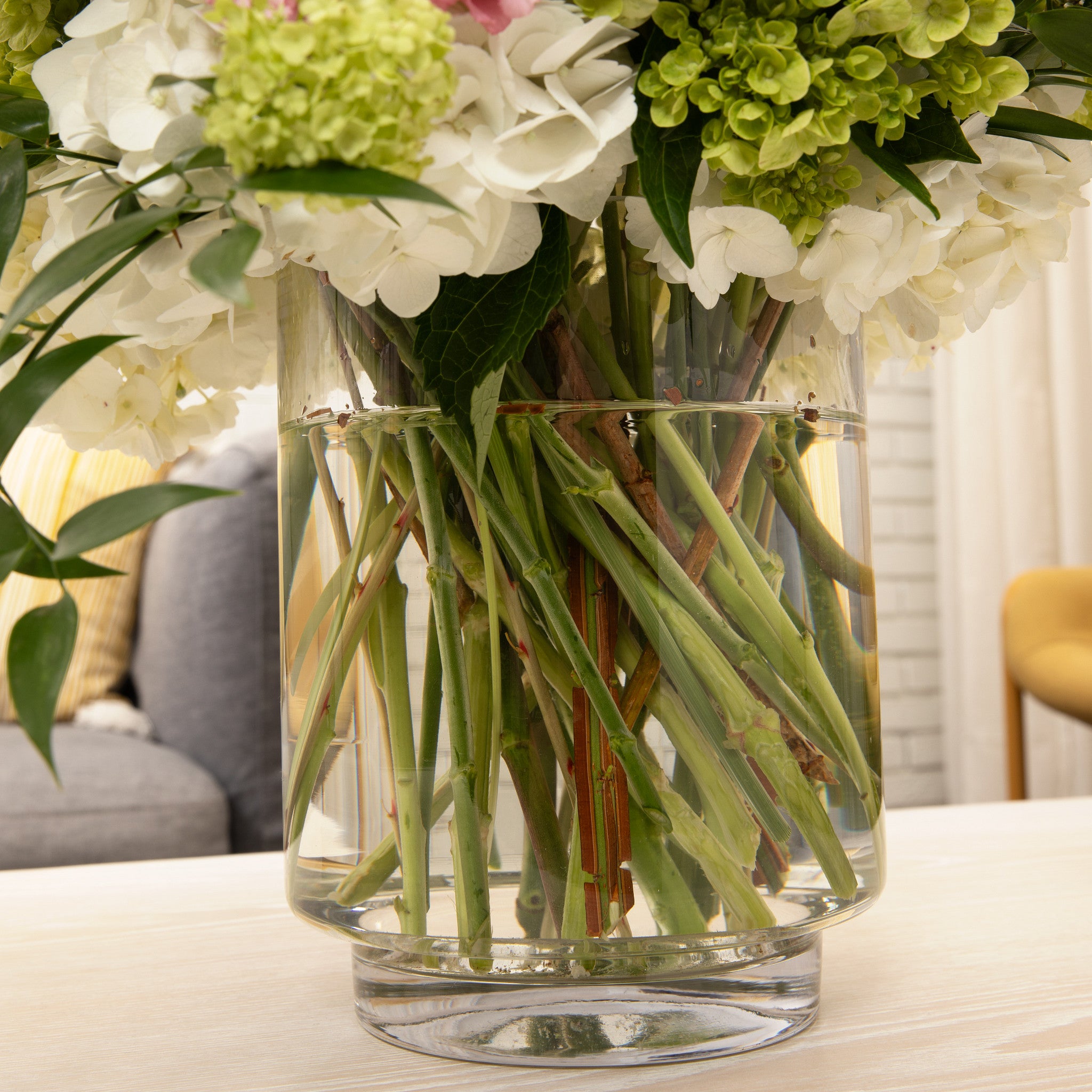 A clear cylindrical glass vase filled with water and fresh cut flowers, including white and light green hydrangeas, set on a light wood table in a softly blurred indoor setting.