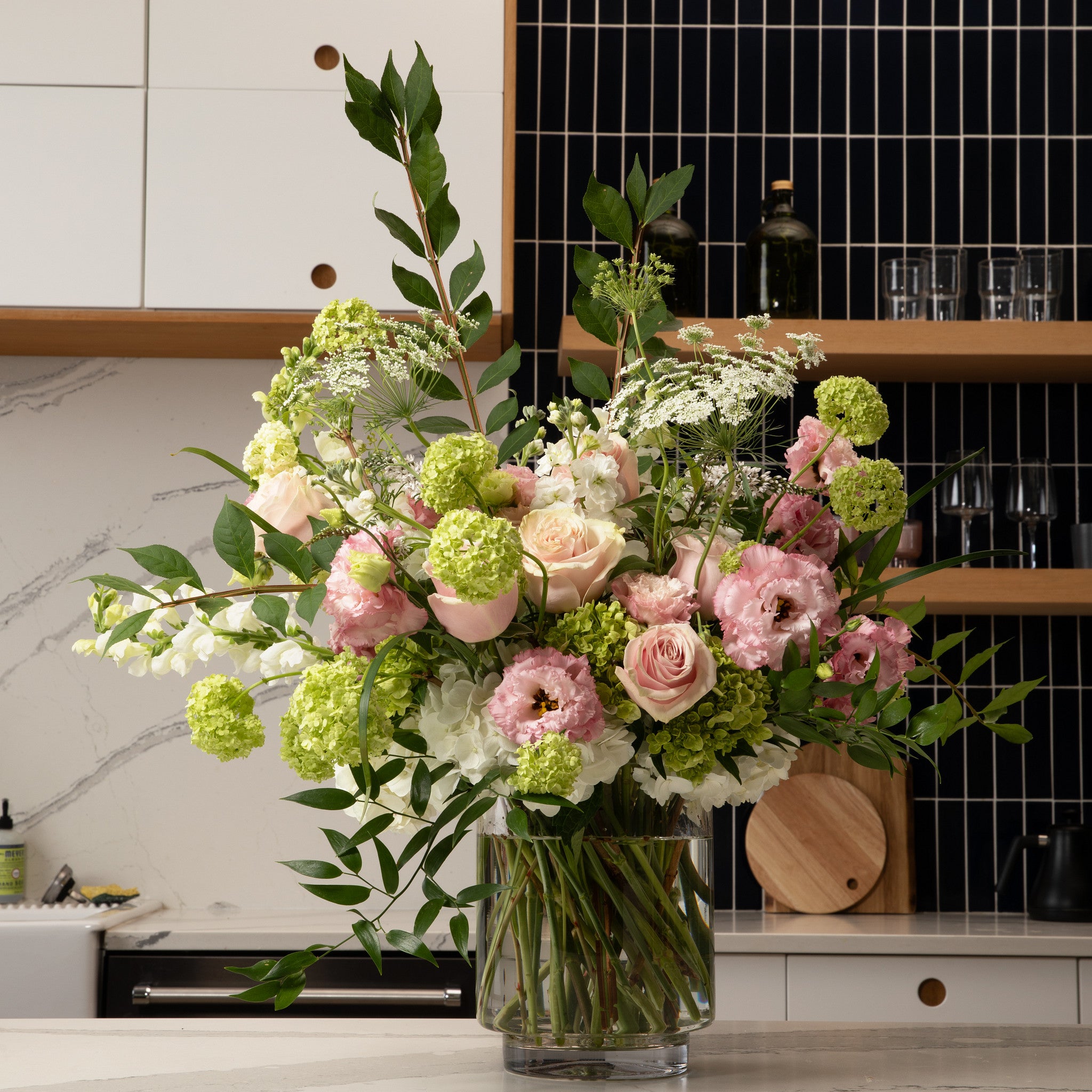 A large, vibrant floral arrangement in a clear glass vase, featuring pink roses, light green hydrangeas, and white flowers, set on a kitchen counter with a modern tiled backsplash.