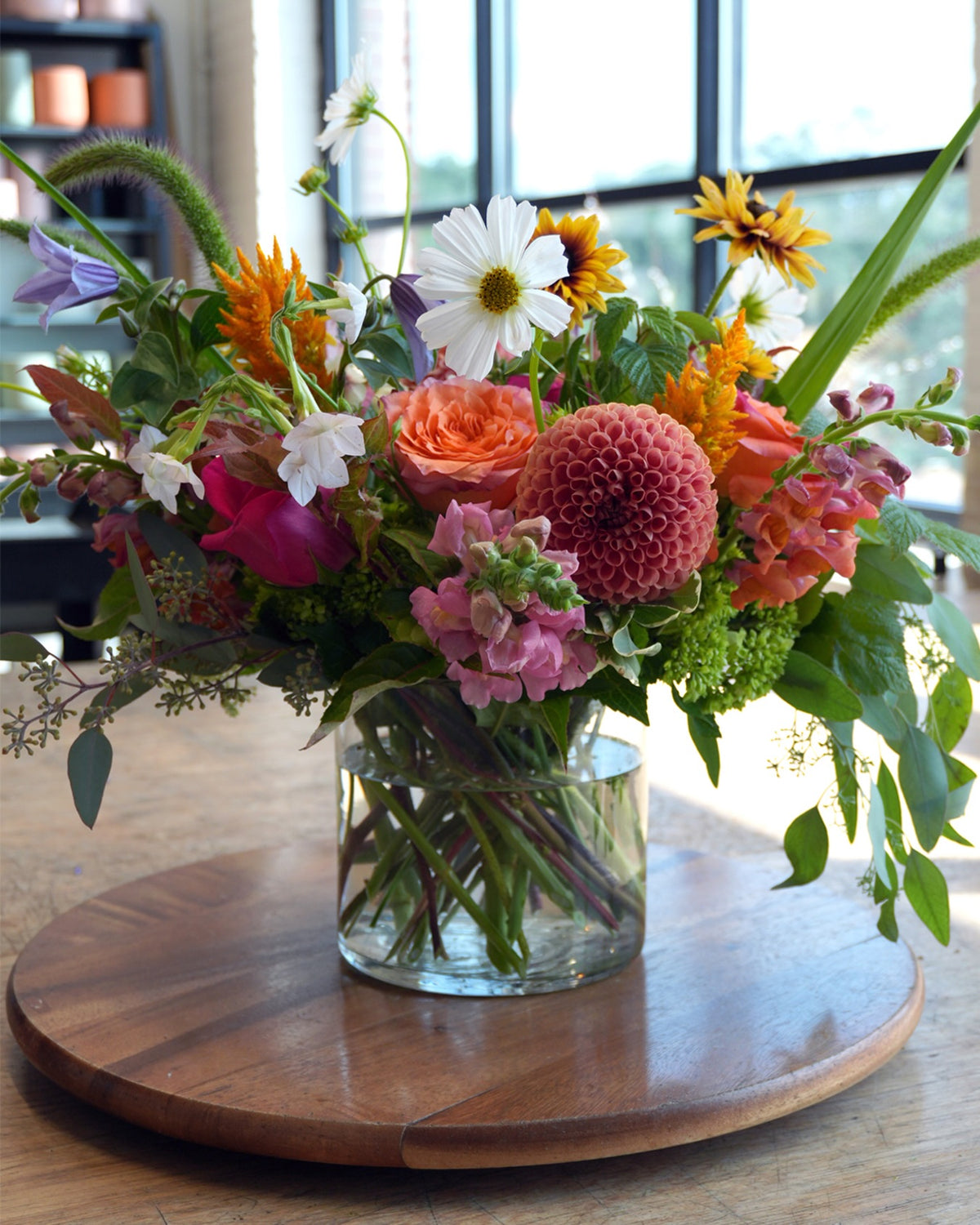A vibrant mixed flower bouquet in a clear glass vase, displayed on a round wooden serving board. The bouquet features a variety of colorful flowers including pink roses, orange dahlias, white daisies, and yellow sunflowers, set against a softly blurred indoor background with a window.