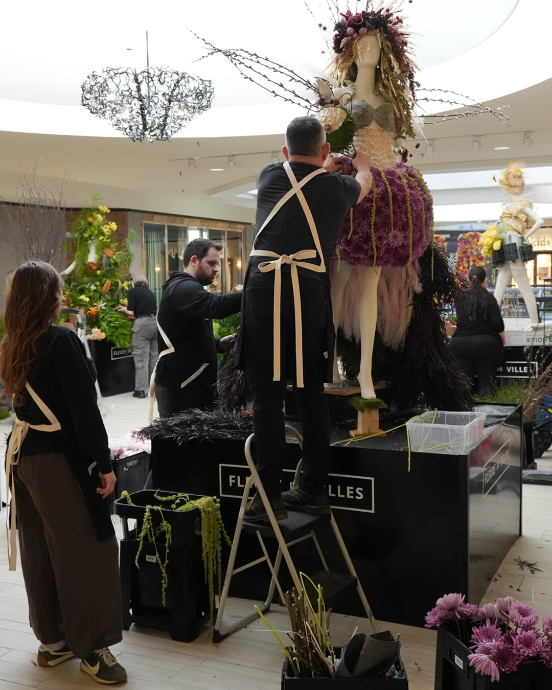 A man in a black apron stands on a step ladder arranging floral decor on a mannequin in a shopping mall atrium. Other people in matching aprons are visible nearby, along with floral arrangements and display tables.