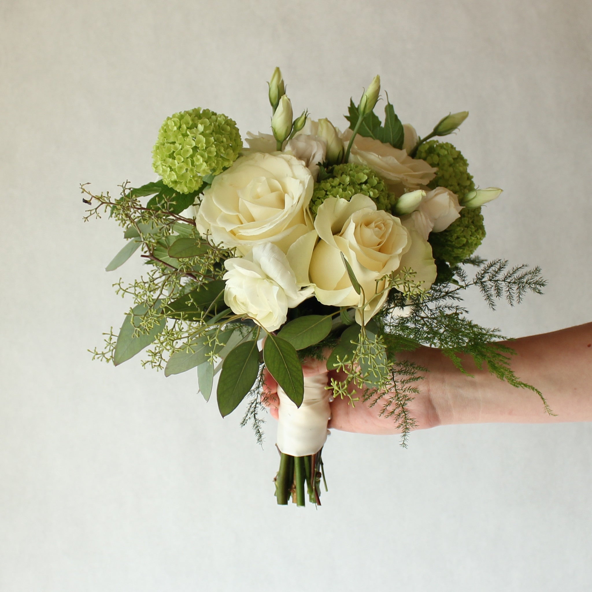 A hand holding a compact bridal bouquet featuring white roses, greenery, and other seasonal blooms, with a  ribbon wrap .